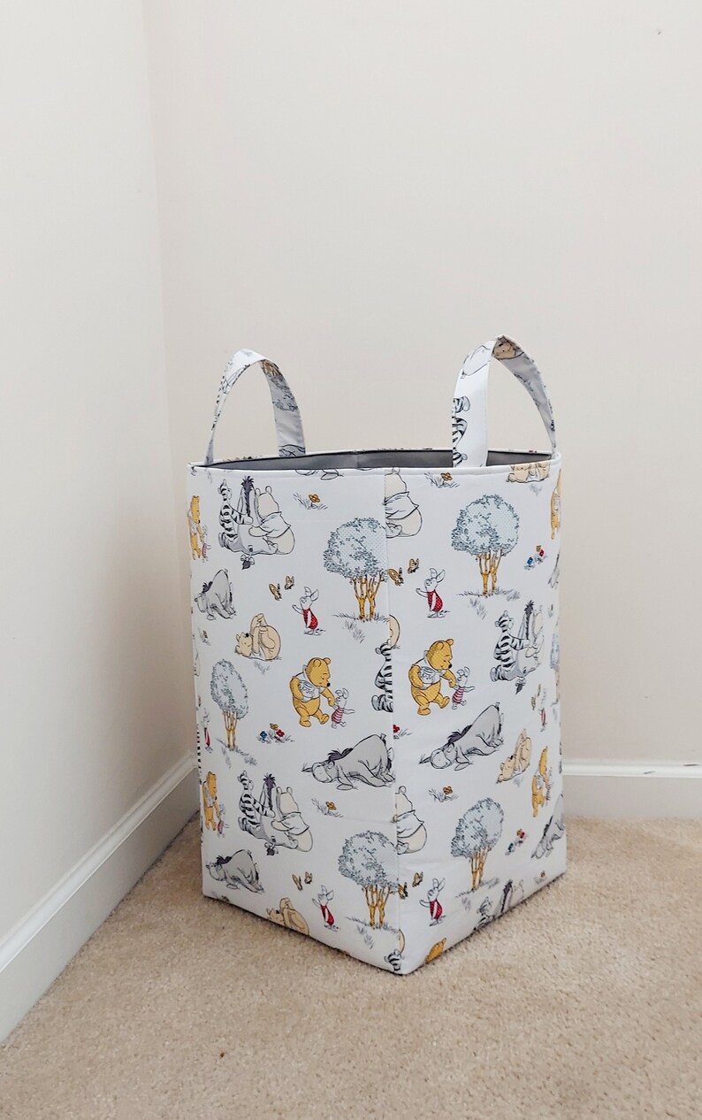 May include: White canvas laundry basket with two handles. The basket is decorated with a Winnie the Pooh print featuring Winnie the Pooh, Piglet, Tigger, Eeyore, and other characters from the Hundred Acre Wood.