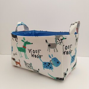 May include: A white fabric storage basket with blue handles and a blue lining. The basket is decorated with a pattern of colorful dogs and the words "Woof Woof".