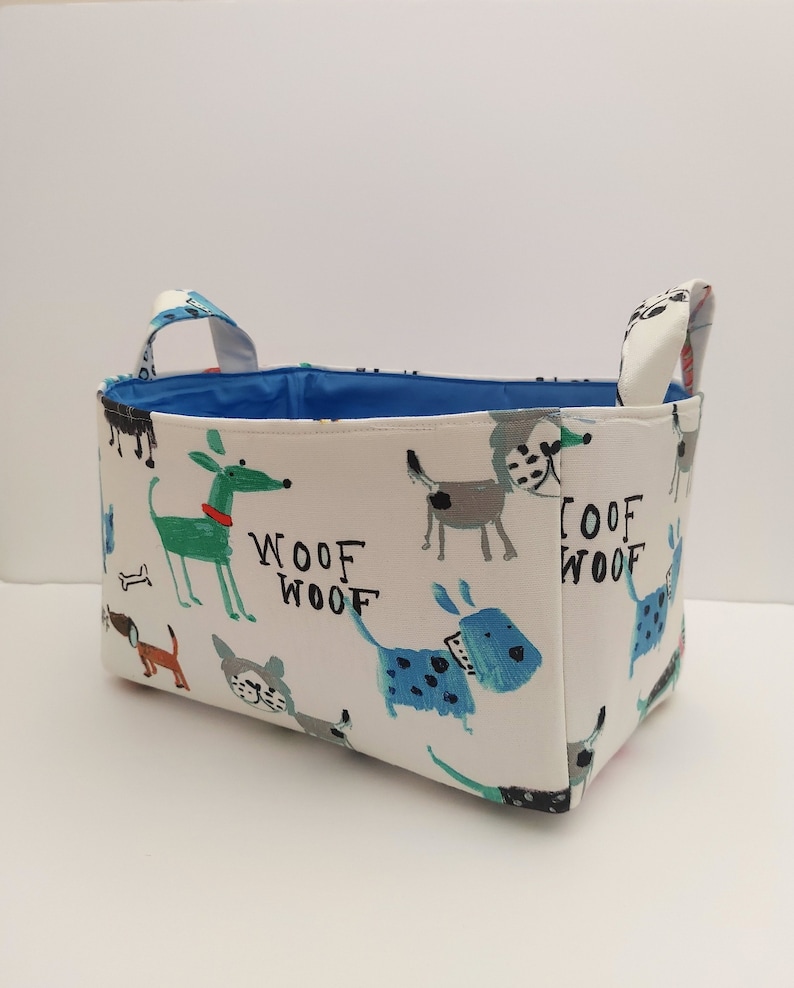 May include: A white fabric storage basket with blue handles and a blue lining. The basket is decorated with a pattern of colorful dogs and the words "Woof Woof".