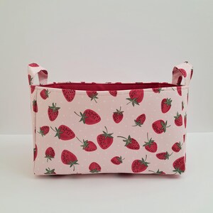 May include: A pink fabric storage bin with red strawberry print and white polka dots. The bin has two fabric handles on the sides.