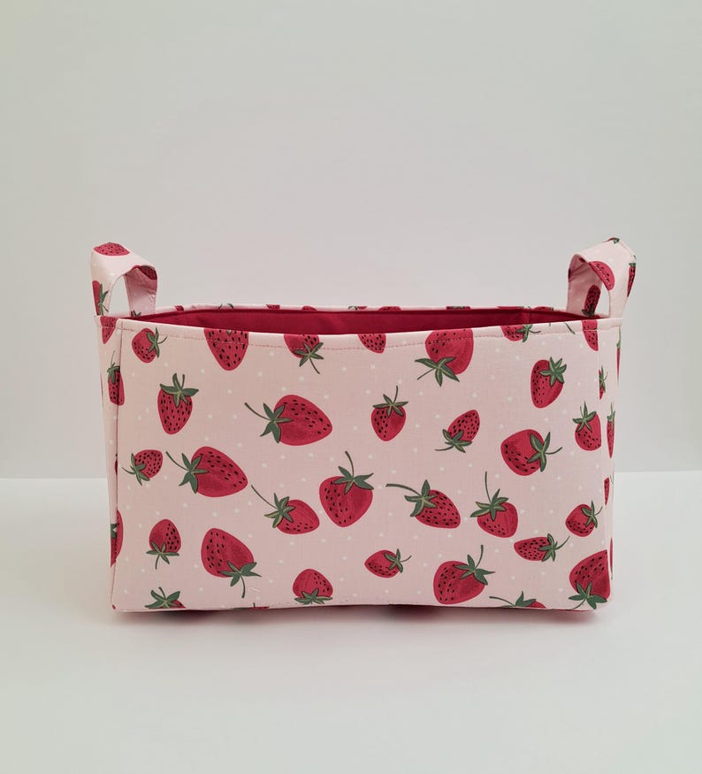 May include: A pink fabric storage bin with red strawberry print and white polka dots. The bin has two fabric handles on the sides.