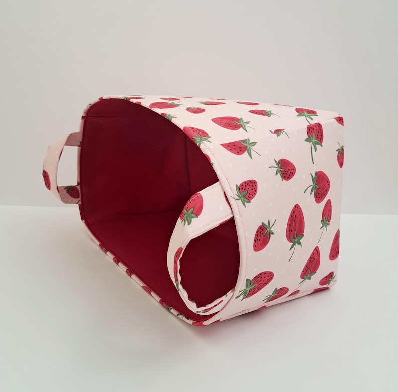 May include: A pink fabric storage basket with a red lining and a strawberry print. The basket has a single handle on one side.