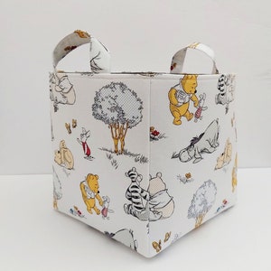 May include: A white fabric storage bin with Winnie the Pooh and friends characters printed on it. The bin has two handles and is square shaped.