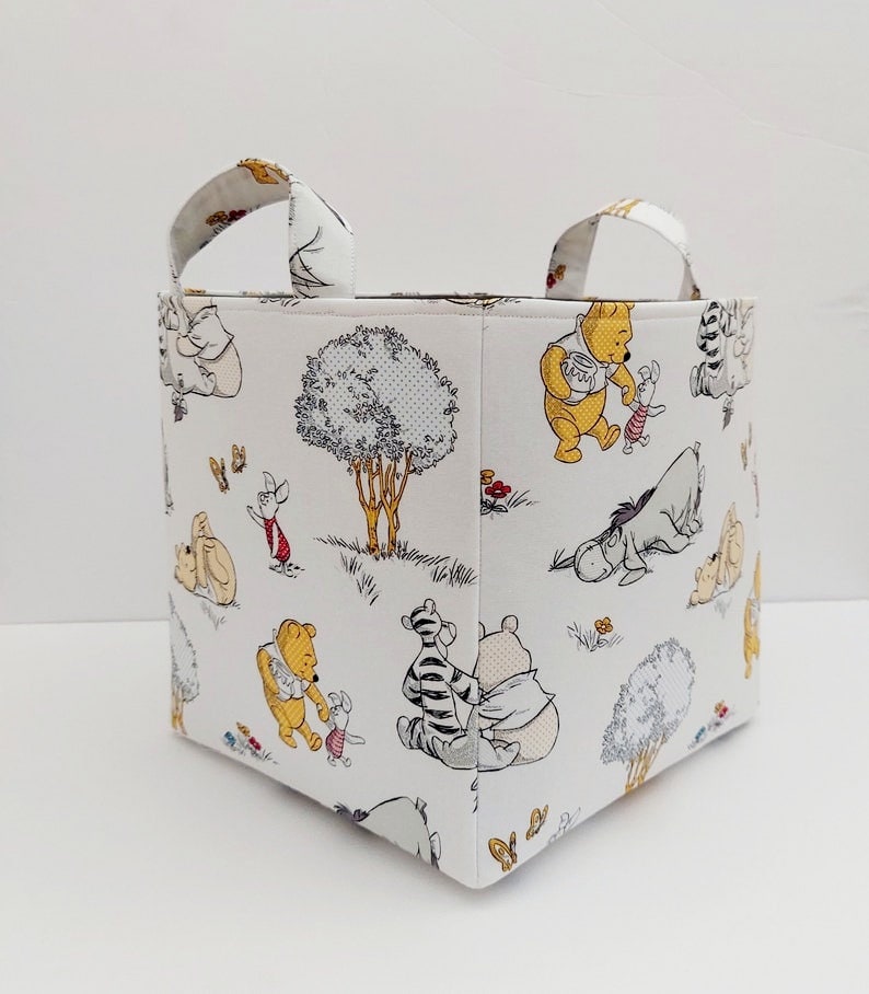 May include: A white fabric storage bin with Winnie the Pooh and friends characters printed on it. The bin has two handles and is square shaped.