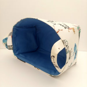 May include: A white fabric storage basket with a blue lining and a handle. The basket is decorated with a whimsical pattern of animals and the words "What a Bow".