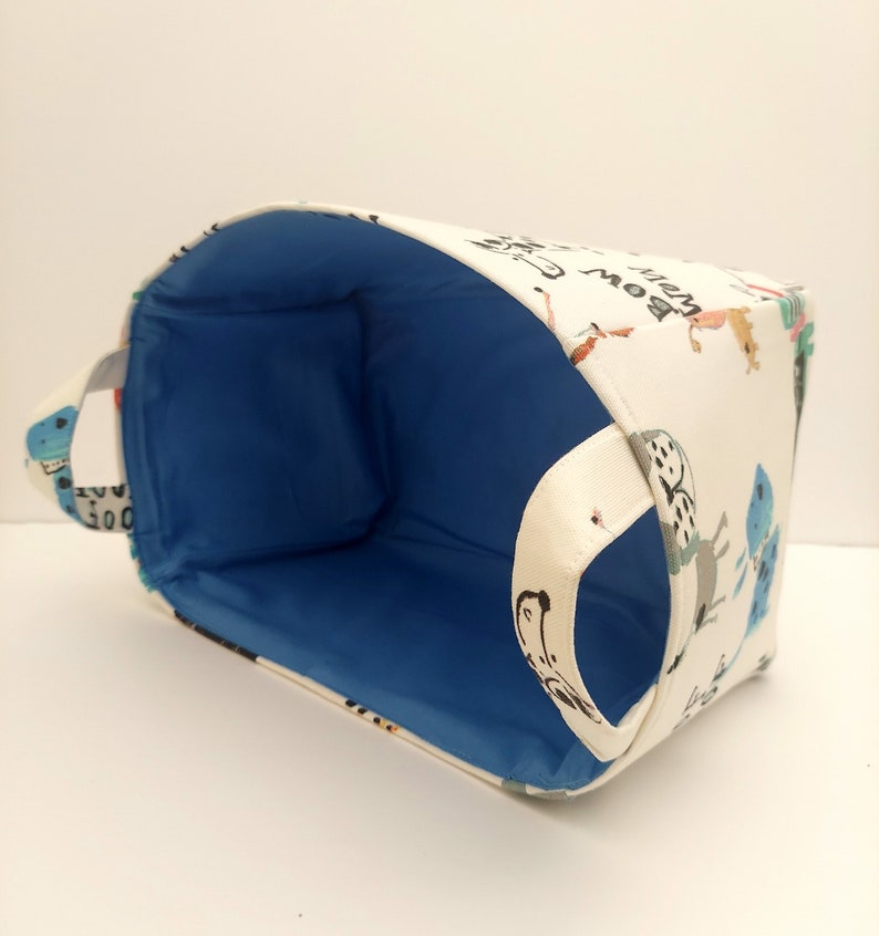 May include: A white fabric storage basket with a blue lining and a handle. The basket is decorated with a whimsical pattern of animals and the words "What a Bow".