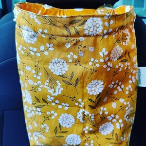 May include: A yellow fabric car trash bag with a white floral pattern. The bag is hanging on the back of a car seat.
