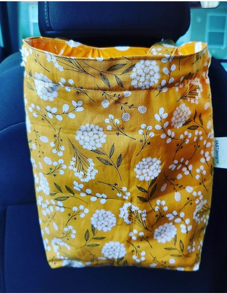 May include: A yellow fabric car trash bag with a white floral pattern. The bag is hanging on the back of a car seat.