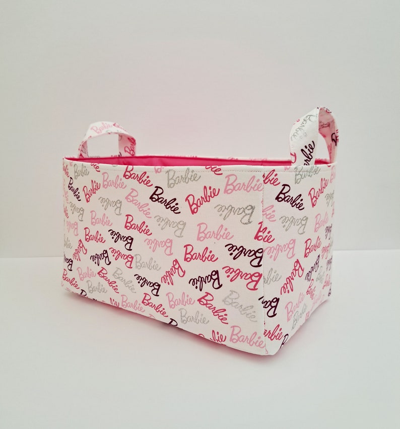 May include: A white fabric storage bin with pink and silver Barbie logos printed all over. The bin has two pink fabric handles.