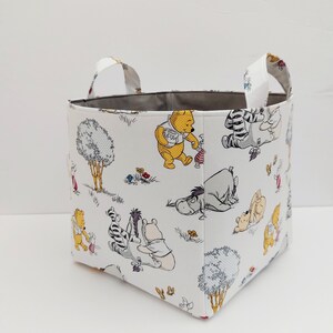 May include: A white fabric storage bin with gray handles. The bin features a repeating pattern of Winnie the Pooh and his friends in a forest setting.