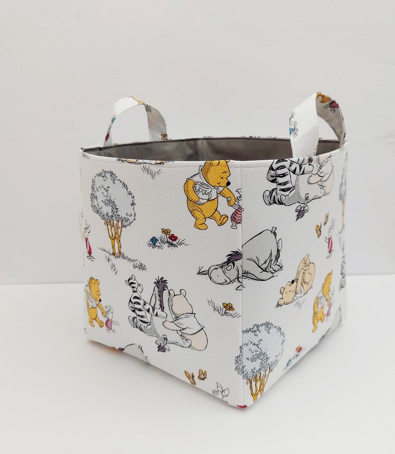 May include: A white fabric storage bin with gray handles. The bin features a repeating pattern of Winnie the Pooh and his friends in a forest setting.