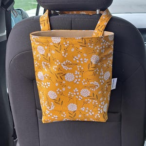 May include: A mustard yellow car seat organizer with a floral pattern of white flowers and brown leaves. The organizer hangs from the headrest and has a rectangular shape with a beige interior. The fabric is a cotton blend.