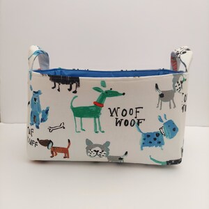 May include: A white fabric storage bin with blue handles and a blue lining. The bin is decorated with a colorful pattern of dogs and the words "Woof Woof".