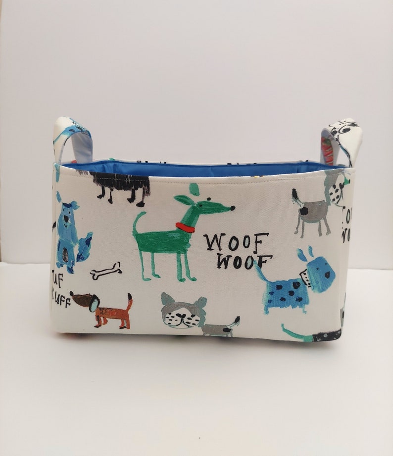 May include: A white fabric storage bin with blue handles and a blue lining. The bin is decorated with a colorful pattern of dogs and the words "Woof Woof".