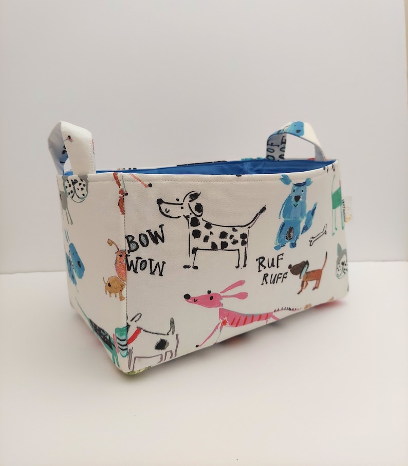 May include: A white fabric storage bin with blue handles. The bin is printed with colorful cartoon dogs and the words 'Bow Wow', 'Ruf Ruff'.