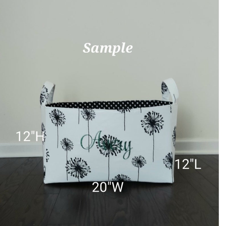 May include: White fabric storage bin with black and white dandelion print and black polka dot lining. The bin has two handles and measures 12 inches high, 20 inches wide, and 12 inches long. The bin is personalized with the name "Amy" in green embroidery.