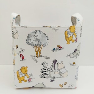 May include: A white fabric storage bin with a Winnie the Pooh and friends print. The bin has two fabric handles on the top and features a repeating pattern of Winnie the Pooh, Piglet, Tigger, Eeyore, and a tree.
