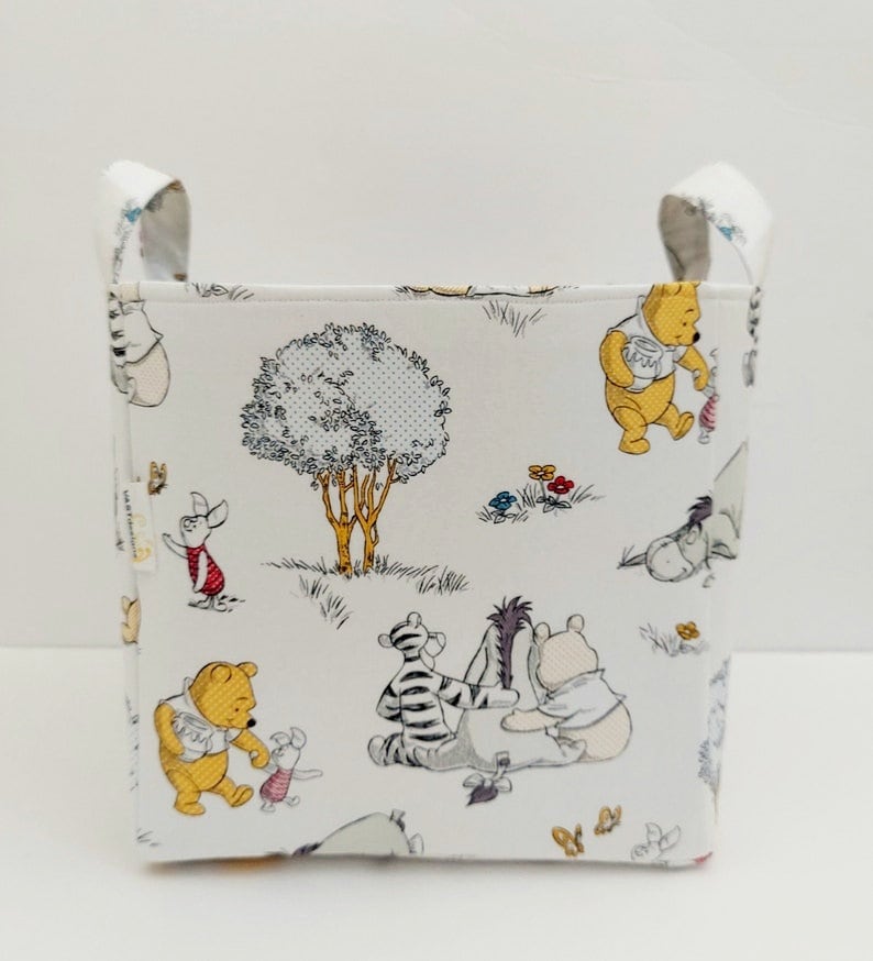 May include: A white fabric storage bin with a Winnie the Pooh and friends print. The bin has two fabric handles on the top and features a repeating pattern of Winnie the Pooh, Piglet, Tigger, Eeyore, and a tree.