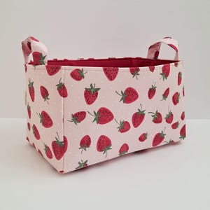 May include: A pink fabric storage bin with red strawberry print and white polka dots. The bin has two handles and is made of a soft, durable fabric.