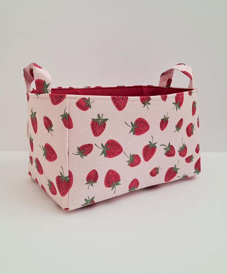 May include: A pink fabric storage bin with red strawberry print and white polka dots. The bin has two handles and is made of a soft, durable fabric.