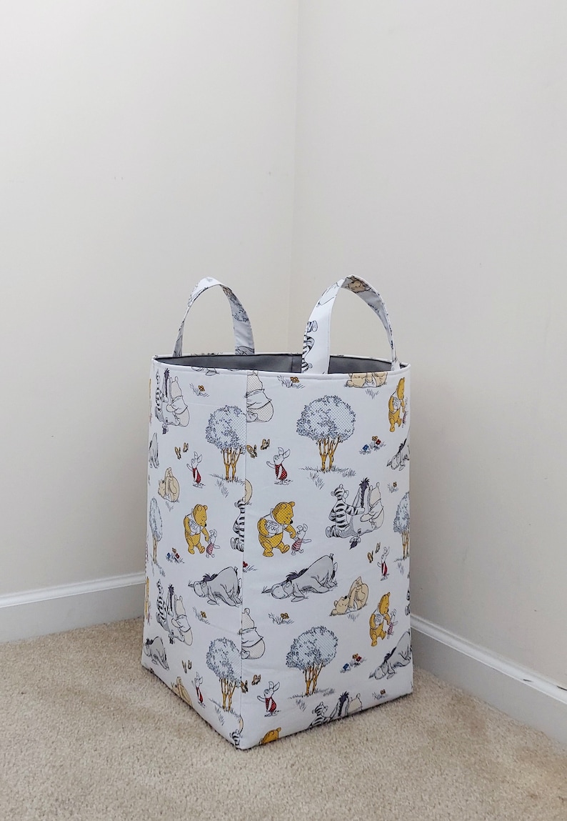 May include: A white fabric laundry basket with Winnie the Pooh and friends printed on it. The basket has two handles and is standing on a beige carpet.
