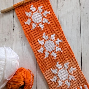 Puede incluir: Un tapiz de pared de ganchillo naranja y blanco con un diseño de sol. La pieza hecha a mano está unida a una clavija de madera y tiene flecos en la parte inferior. Dos ovillos de lana, uno blanco y otro naranja, están junto al tapiz.