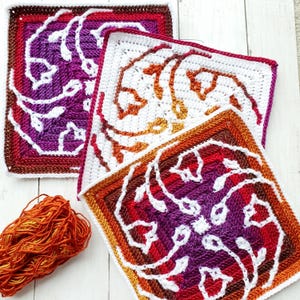 May include: Three square crochet pieces with a floral design in shades of purple, red, orange, and white. A ball of orange yarn sits to the left of the squares. The crochet pieces are on a white wooden surface.