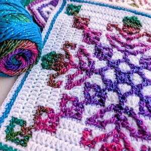 May include: A close-up of a white crocheted square with a snowflake design in shades of purple, pink, and green. A roll of blue and multicolored yarn sits next to the square. The square has a teal border.