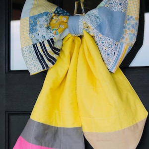 May include: A large decorative bow made of fabric, featuring a patchwork pattern of blue, yellow, and white floral and striped prints on the top section. The two long hanging tails are solid yellow with grey, pink, and tan color-blocked ends.