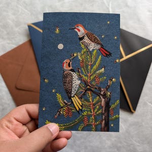 May include: A blue card with a painting of two red-breasted woodpeckers perched on a pine tree branch. The birds are surrounded by fireflies and a full moon.