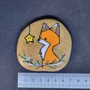 Op de afbeelding: Handgeschilderde steen met een vos met oranje vacht en een witte borst, die omhoog kijkt naar een gele ster die aan een touwtje hangt. De steen is versierd met groene bladeren en rode bessen.