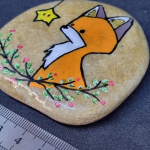 Op de afbeelding: Handgeschilderde steen met een oranje vos met een gele ster erboven. De vos is omringd door groene bladeren en roze bessen.