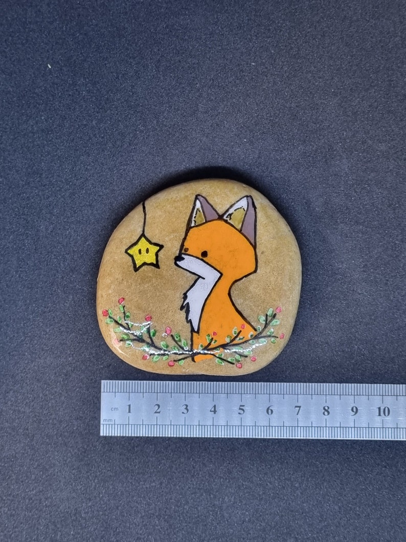 Op de afbeelding: Handgeschilderde steen met een vos met oranje vacht en een witte borst, die omhoog kijkt naar een gele ster die aan een touwtje hangt. De steen is versierd met groene bladeren en rode bessen.