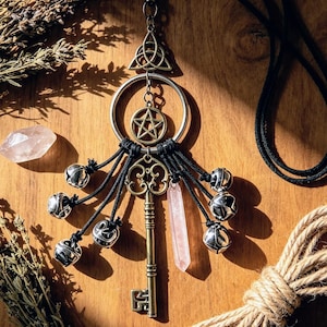 May include: A decorative wall hanging featuring a brass-toned skeleton key, a pentagram charm, and a triquetra symbol suspended from a metal ring. Six silver-toned jingle bells and a pale pink crystal point hang from black cords attached to the ring.
