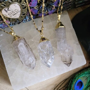 May include: Three necklaces featuring clear, raw crystal points suspended from gold-toned metal chains. Each crystal is capped with a gold-toned metal bail. The pendants are arranged vertically, showcasing the natural, jagged texture of the stones.