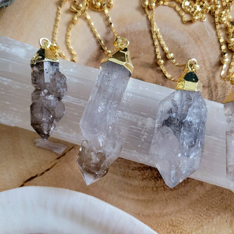 May include: Three necklaces featuring raw, translucent, smoky quartz crystal points suspended from gold-toned metal caps and delicate gold-toned chains. Each crystal has a unique, jagged, natural shape with varying shades of gray and clear quartz.