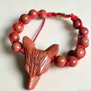 May include: A bracelet featuring a carved red stone fox head pendant and round red stone beads strung on a bright red cord. The design includes a small gold-toned metal accent piece near the top of the cord, connecting the beaded sections.