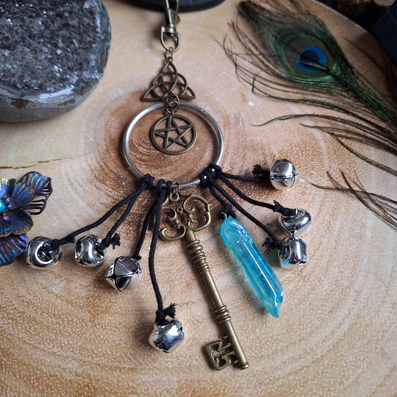 Puede incluir: Un cristal azul, una llave y un amuleto de pentagrama cuelgan de un anillo plateado con cordones negros y cascabeles plateados. Los amuletos est&aacute;n unidos a un cord&oacute;n negro con un nudo en la parte superior.