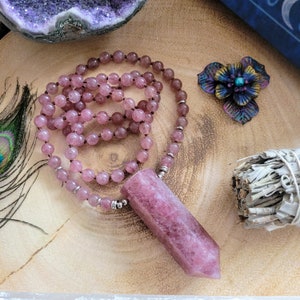 May include: A pink gemstone pendant necklace with a beaded strand. The necklace features a large, rectangular pendant and small, round beads. A decorative metal flower and a bundle of sage are also visible.