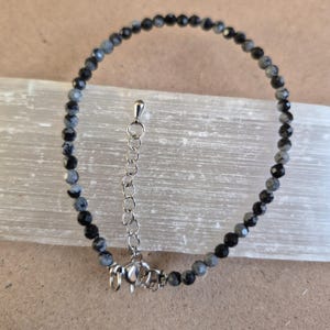 May include: A delicate bracelet featuring small, faceted, round beads in alternating shades of charcoal grey and black. The jewellery piece includes a silver-toned metal extender chain with a lobster claw clasp, allowing for adjustable sizing.