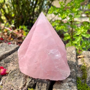 May include: A large, pale pink rose quartz crystal shaped like a pyramid. The crystal is sitting on a rough, gray stone surface.
