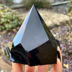 May include: A black obsidian pyramid crystal, a type of volcanic glass, with a smooth, polished surface.