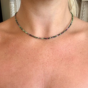 May include: A finely crafted necklace featuring tiny beads in shades of green, red, and black. The beads are strung together to create a choker-style necklace. The necklace is shown around a person's neck.