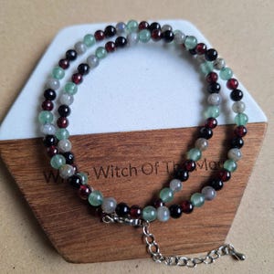 May include: A beaded necklace with green, red, and black gemstones. The necklace is displayed on a wooden and white geometric stand. The necklace has a silver clasp and extender chain.