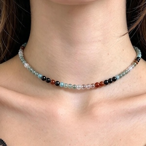 May include: A beaded choker necklace featuring a sequence of small, polished spherical beads in various colours including translucent light blue, opaque black, deep reddish-brown, and clear white, arranged in a repeating pattern around the neck.