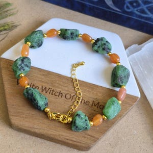 May include: A bracelet featuring alternating raw, green and black speckled stone nuggets and smooth, oval orange beads. The stones are separated by small, round gold-toned metal beads. The bracelet includes a gold-toned metal chain and lobster clasp.