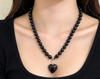 May include: A necklace featuring a strand of smooth, round, polished black beads. A single, glossy black heart-shaped pendant hangs from a small silver-toned metal bail at the center of the bead strand, resting against a black shirt.