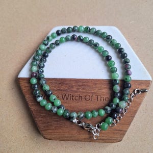 May include: A necklace made of green and black beads, with some red beads. The necklace is presented on a hexagonal coaster made of wood and white material. The text "The Witch Of The" is visible.