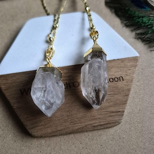 May include: Two clear, raw quartz crystal pendants suspended from gold-toned metal chains. Each crystal is capped with a gold-toned metal setting at the top, featuring a faceted, pointed shape that hangs vertically from the delicate link chains.