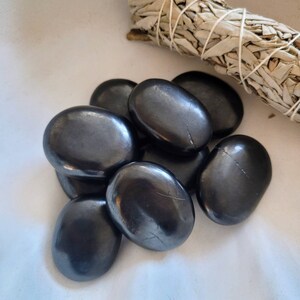 May include: A group of eight smooth, black obsidian stones, also known as black glass, in various sizes and shapes. The stones are arranged in a pile on a white surface.